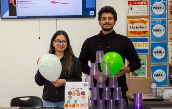 Volunteer lead and a student each hold balloons and stand behind a table with a full tower of plastic cups