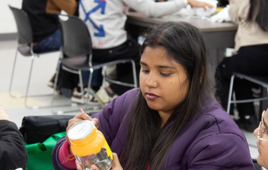 A student in a purple jacket sits at a table painting a jar 