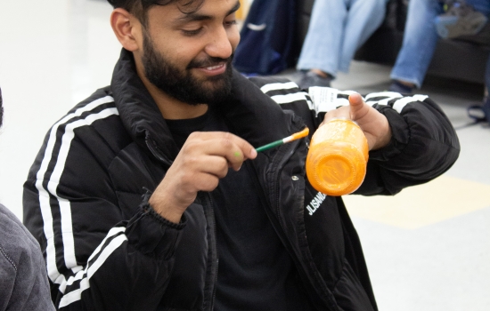 A student wearing a black track jacket sits at a table painting a jar 