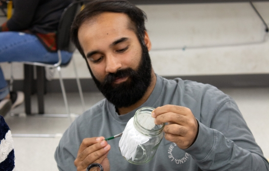 A student with a beard sits at a table painting a jar 