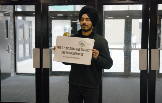 Pratham holds up a sign that says "Find 3 people wearing glasses and bring them here"