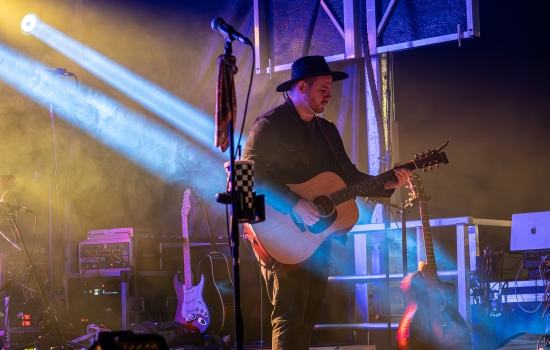 A musician with an acoustine guitar and a hat performs on a stage 