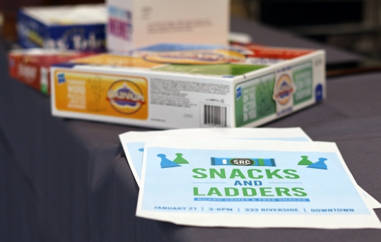 The Snacks and Ladders event flyer sits on a table with a snack of board games in the background