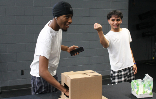 A student feels inside a mystery box while another watches and encourages