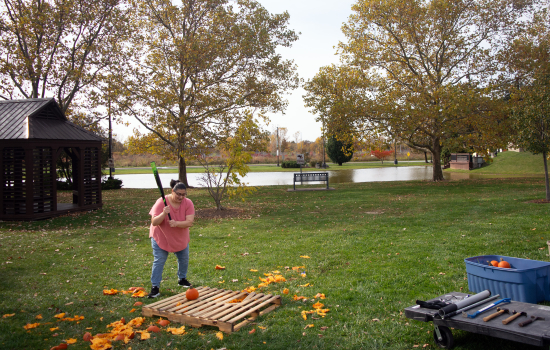 Action shot of a student preparing to smash a pumpkin with a baseball bat outside on the grass in front of the pond