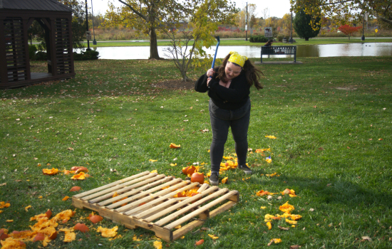 Action shot of a student smashing a pumpkin with a crowbar outside on the grass in front of the pond